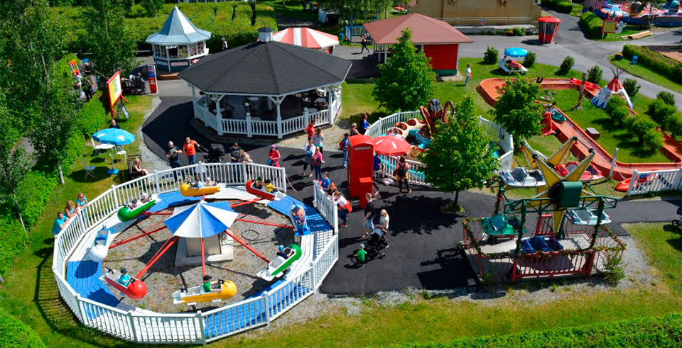 Ilmakuva pienestä, värikkäästä huvipuistosta, jossa näkyy lapsille suunnattuja huvilaitteita ja vihreää kesäistä ympäristöä.