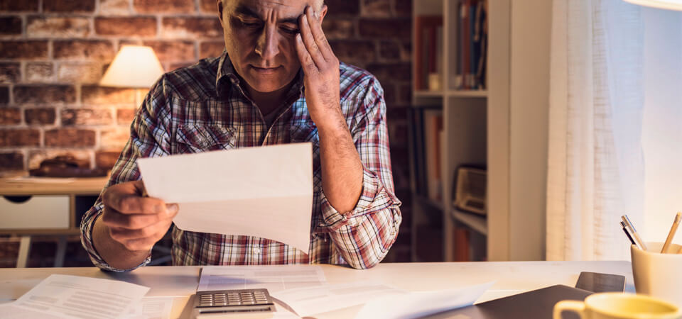 Huolestunut mies lukee kotona laskua; pöydällä laskin ja paperipino – esimerkki tilapäisestä rahantarpeesta.